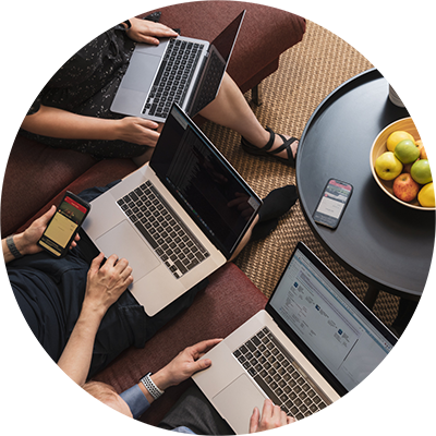 Three people sitting on a sofa with laptop computers on their laps from a top view. The person in the middle holds a smartphone in their hand. In front of the people is a sofa table with another smartphone and a fruit bowl.